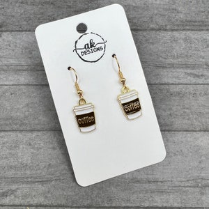 May include: Gold-coloured dangle earrings with a coffee cup design. The earrings have a white cup with a brown band and the word "coffee" in a brown font. The earrings are displayed on a white card with the "ak designs" logo.