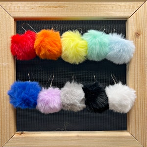 May include: Assortment of 10 colorful faux fur pom-pom earrings. The earrings are displayed on a black mesh background. The pom-poms are in the colors red, orange, yellow, light blue, blue, lavender, gray, black, and white.