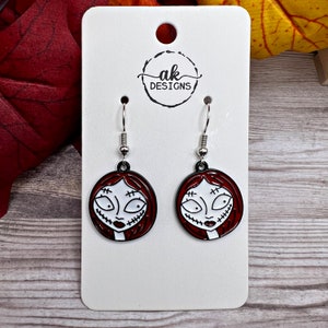 May include: A pair of silver dangle earrings with red and white enamel charms featuring a cartoon character with stitched-up eyes and mouth. The charms are round and have a black outline. The earrings are on a white card with the text "ak DESIGNS".