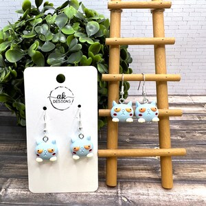 May include: A pair of blue cat dangle earrings with white eyes and orange eyebrows. The earrings are hanging from a white card with the text "ak DESIGNS" printed on it.