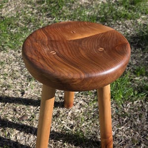 May include: A handcrafted wooden stool with a round, dark brown seat and three light brown legs. The seat has a natural wood grain pattern and is secured with small wooden pegs. The stool is set on a grassy surface.