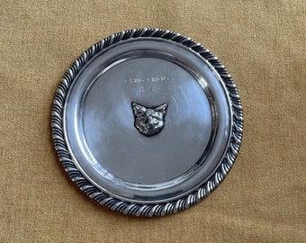 Puede incluir: Un plato redondo decorativo de color plateado con un diseño de cabeza de zorro en relieve en el centro. El plato tiene un borde con cuentas y el texto "The Fox Troop 11.58" está grabado en la superficie.