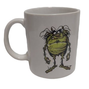 May include: White ceramic coffee mug with a cartoon illustration of a green, hairy creature with large eyes and a toothy grin.