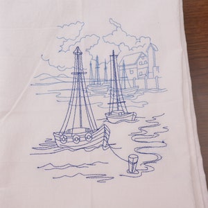 Puede incluir: Toalla de cocina blanca bordada con un diseño azul de veleros en un puerto. El diseño incluye un pequeño edificio, nubes y agua.