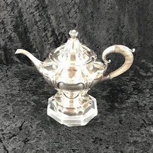 May include: A silver teapot with an ornate design and a curved spout. The teapot is sitting on a clear glass pedestal.