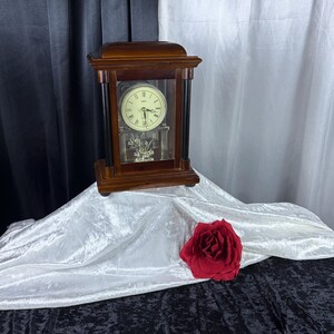May include: A rectangular wooden mantel clock with a dark brown finish and black pillars. The clock face is cream-colored with black Roman numerals and the brand name Seiko. A silver-toned pendulum is visible behind the glass front panel.
