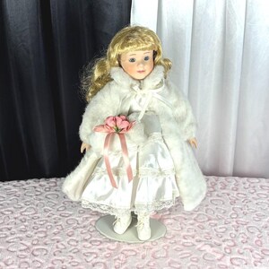 May include: A porcelain doll with blonde hair and blue eyes, wearing a white satin dress with lace trim and a white faux fur coat. The doll holds a small white muff decorated with a pink flower and ribbon, and wears white lace-up boots.