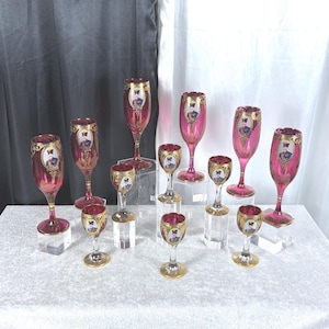 May include: A collection of eleven cranberry-colored glass goblets and stems featuring gold-rimmed oval portraits of historical figures. Each piece has a clear glass base and stem, with ornate gold filigree detailing surrounding the portrait.