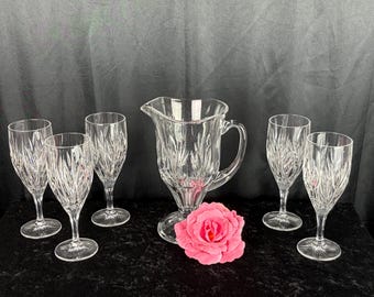 May include: A clear glass pitcher and four matching stemmed glasses, each featuring a vertical cut-glass pattern. A single vibrant pink rose is positioned in front of the pitcher, which has a curved handle and a wide pouring spout.