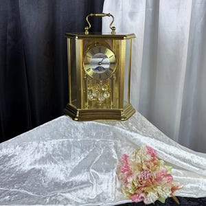 May include: A gold-tone mantel clock with a glass-paneled case, a top carrying handle, and a circular face featuring Roman numerals. The clock includes a moon phase indicator and a rotating pendulum with clear crystal spheres at the base.