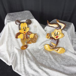 May include: Two handcrafted wooden wall plaques featuring cartoon characters. One plaque depicts a mouse figure in a brown jacket holding a suitcase, while the other shows a rabbit figure with long ears holding a small rectangular object.