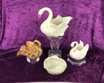 May include: A collection of four swan-shaped decorative vessels. One large white swan sits on a clear glass pedestal, while a smaller white swan, a gold-toned swan, and a small white swan-shaped bowl rest on clear glass bases.