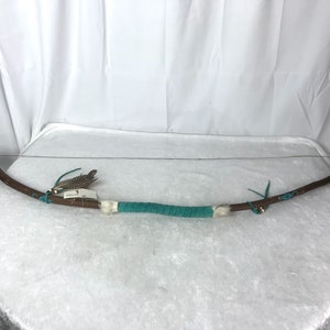 May include: A wooden bow with turquoise and white accents. The bow is strung with a thin string and has feathers and bells attached to the ends.