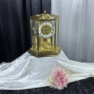 May include: A gold-toned mantel clock with a hexagonal glass case, featuring a white dial with black Roman numerals and a central gold filigree pattern. The clock includes a rotating pendulum with four gold spheres and etched floral glass panels.
