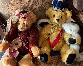 May include: Two plush teddy bears. The left bear wears a deep red coat, a floral headpiece, and a decorative mask. The right bear wears a blue velvet crown, a red sash with a gold emblem, and a white faux fur cape with gold rope detailing.