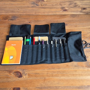 Peut inclure: Organisateur de fournitures d'art enroulable en toile noire avec plusieurs poches et rabats. Contient diverses fournitures d'art, dont des crayons de couleur, des stylos et un carnet avec le mot "Expose" sur la couverture. Ciseaux visibles à droite.