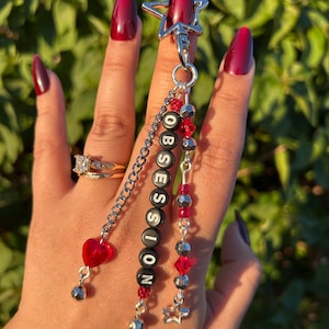May include: A silver star-shaped keychain featuring three dangling strands. One strand has a red heart bead, another has black letter beads spelling OBSESSION, and the third features alternating red and silver beads with a small silver star charm.