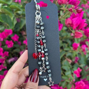 May include: A silver-toned keychain charm featuring multiple dangling chains attached to a lobster clasp. The chains are adorned with black letter beads spelling out "I LOVE SO SO MUCH," interspersed with red faceted beads, silver beads, and star charms.