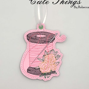 May include: A pink fabric ornament shaped like a spool of thread with a needle, featuring a floral design. The ornament has the text "Cute Things by Rebecca" in black script and is suspended by a white ribbon.