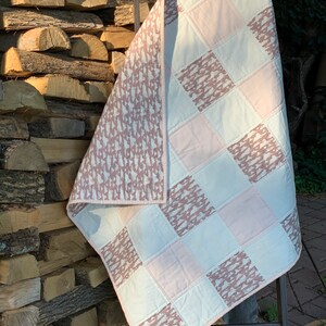 May include: A pink and white patchwork quilt with a brown and white patterned design. The quilt is folded and leaning against a stack of firewood.