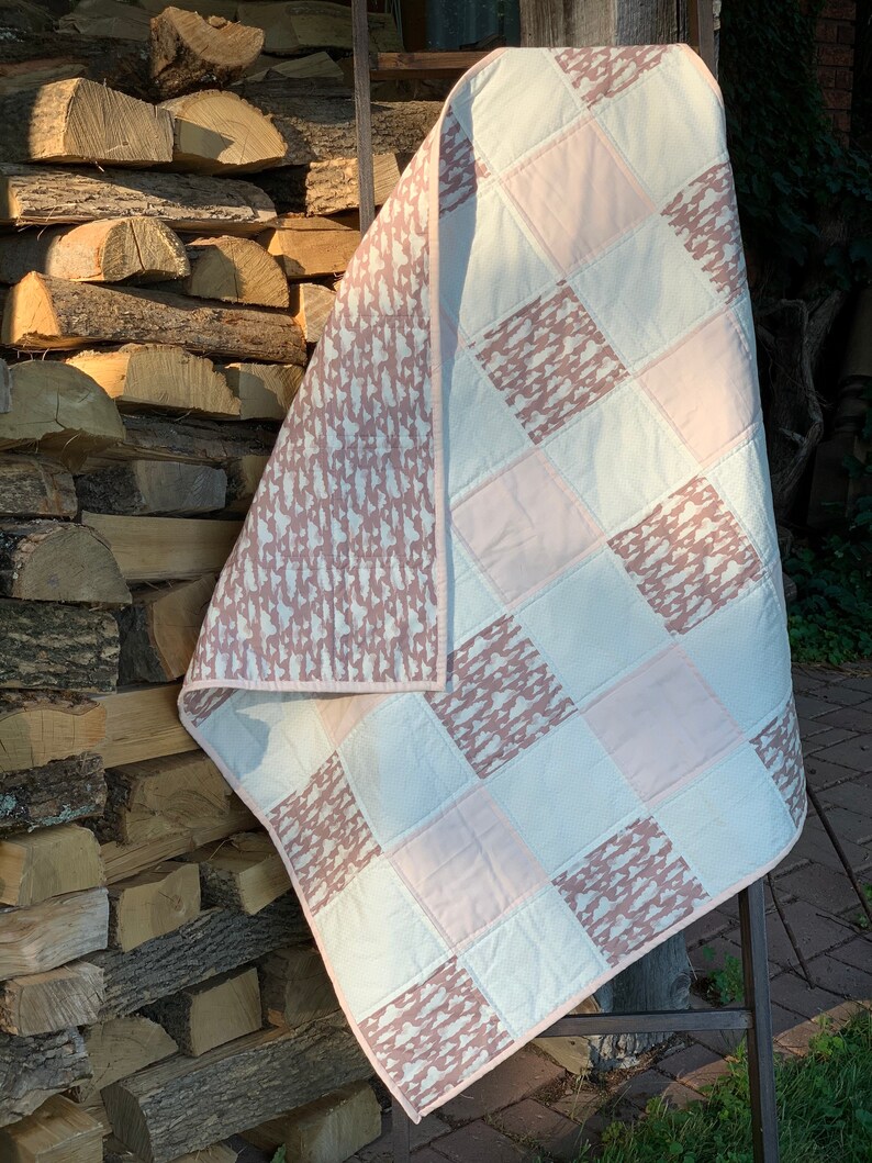 May include: A pink and white patchwork quilt with a brown and white patterned design. The quilt is folded and leaning against a stack of firewood.