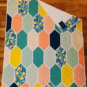 May include: A quilt with a geometric pattern of hexagons in shades of pink, yellow, green, and blue. The hexagons are outlined in white and the quilt is backed with white fabric. The quilt has a label that says "Lissy".
