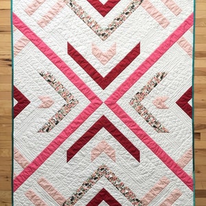 May include: A white quilt with a geometric pattern of pink, red, and floral fabric. The quilt is edged with a teal border.
