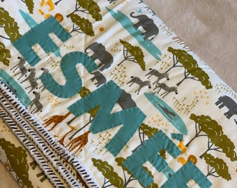 May include: A white and green baby blanket with a safari animal print. The blanket has a blue embroidered name "Esme" on it.