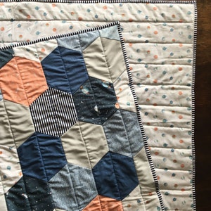 May include: A quilt with a geometric pattern of hexagons in shades of blue, gray, and orange. The quilt is made of different fabrics with various patterns, including stripes, polka dots, and a space-themed print. The quilt is edged with a black and white trim.