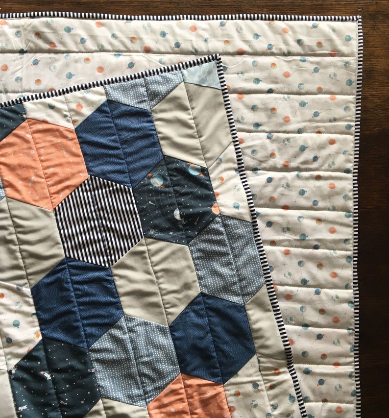 May include: A quilt with a geometric pattern of hexagons in shades of blue, gray, and orange. The quilt is made of different fabrics with various patterns, including stripes, polka dots, and a space-themed print. The quilt is edged with a black and white trim.