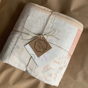 May include: A white and pink patchwork quilt with a floral pattern is wrapped in brown paper and tied with twine. A small brown tag with the text "Fox & Cedar Designs" is attached to the twine. A handwritten note with the text "love from,  [heart]" is attached to the tag.