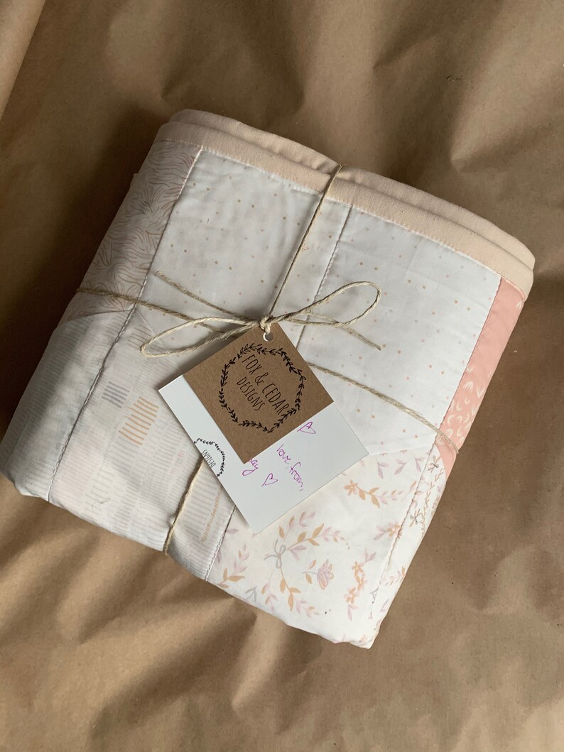 May include: A white and pink patchwork quilt with a floral pattern is wrapped in brown paper and tied with twine. A small brown tag with the text "Fox & Cedar Designs" is attached to the twine. A handwritten note with the text "love from,  [heart]" is attached to the tag.