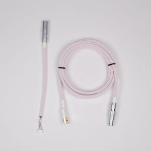 Könnte beinhalten: Aufgerolltes, hellrosa USB-Kabel mit silbernen Steckern und einem weißen USB-A-Anschluss. Das Kabel ist mit einem geflochtenen Stoff umwickelt und mit schwarzen Bindern befestigt. Das Kabel hat einen Stecker mit freiliegenden Drähten.