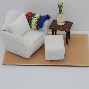 May include: Miniature living room set with a white sofa, ottoman, and a small wooden side table. A colorful crocheted pillow rests on the sofa. A small vase with a white flower sits on the table. The set is on a tan rug.