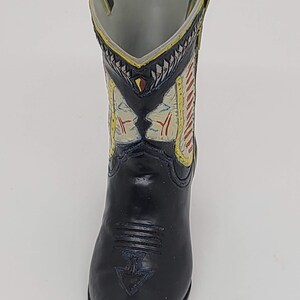 May include: A black cowboy boot-shaped vase with a decorative design. The boot features a pointed toe and a detailed pattern on the upper shaft with red, yellow, and white accents. The interior is gray.