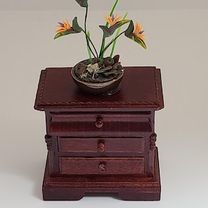 May include: Miniature wooden nightstand with three drawers, a dark reddish-brown finish, and decorative carvings. A small ceramic bowl with a miniature bird of paradise plant sits on top, featuring orange and purple flowers.