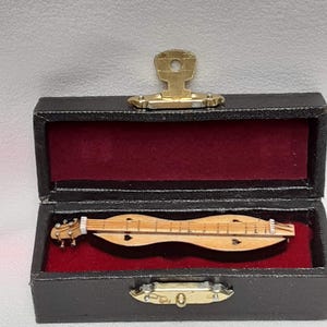 May include: A miniature wooden dulcimer rests inside a black case with a red velvet lining. The case has a gold-colored clasp and hinges. The dulcimer has a light brown finish with strings and decorative heart-shaped sound holes.