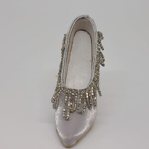 May include: A decorative, miniature silver shoe with a pointed toe. The shoe is embellished with silver beads along the top and around the opening, with beaded fringe cascading down the front. The shoe has a gold-colored sole.