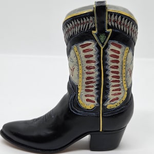 May include: A black cowboy boot figurine with decorative detailing. The boot features a pointed toe, a small heel, and intricate designs in red, yellow, and gray on the shaft. The top of the boot is gray with a yellow trim.