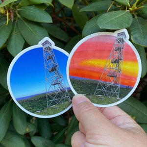 May include: Two round stickers featuring a fire tower illustration. One shows the tower against a blue sky, the other against a sunset. The stickers have a white border and are held against a backdrop of green leaves.