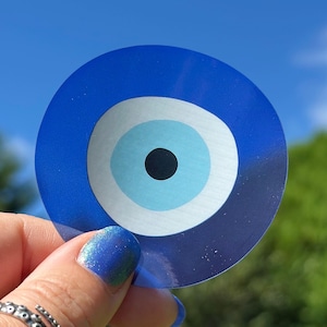 May include: A circular blue Evil Eye sticker featuring a light blue and white iris and a black pupil. The sticker is set against a backdrop of a bright blue sky and green foliage.