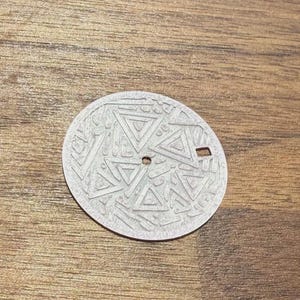 May include: A circular silver-toned watch dial featuring a textured surface with embossed geometric triangle patterns. The dial includes a central hole for hands and a small rectangular date window aperture positioned on the right side of the face.