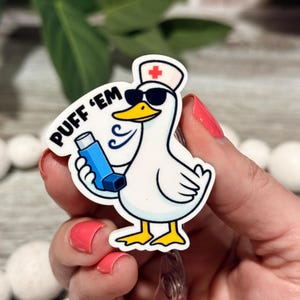 May include: A white duck badge reel featuring a cartoon duck wearing a nurse cap with a red cross and black sunglasses. The duck holds a blue inhaler, with blue vapor swirls and the black text PUFF 'EM printed above the inhaler on the white background.
