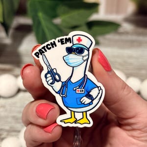 May include: A badge reel featuring a cartoon goose dressed as a medical professional in a blue scrub top, a stethoscope, a face mask, and a nurse cap. The goose wears sunglasses, holds scissors, and has the text PATCH 'EM with a red cross symbol.