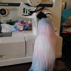 May include: A light blue, pink, and white ombre faux fur tail with a black string attached to it. The tail is hanging from a white sewing machine with a price tag that says "$15".