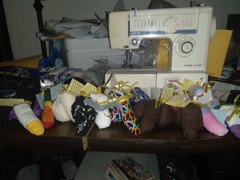 May include: A collection of colorful fabric-covered bean bags with yellow ribbons and tags. The bean bags are arranged on a wooden table in front of a white sewing machine.