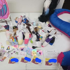 May include: A table top display of colorful handmade sock puppets with various designs and patterns. The puppets are arranged in rows and include a variety of colors, including pink, blue, green, and yellow. There are also several small cards with text on them, including "Back Dot" and "Welcome Home, Mia".