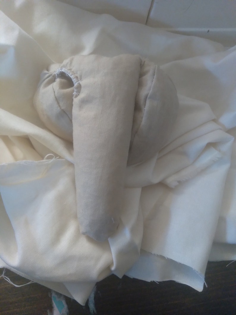 May include: A beige fabric arm for a stuffed animal, with a hole for the head, lying on a white fabric surface.