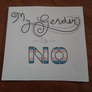 Puede incluir: Una nota adhesiva blanca con escritura cursiva negra que dice "My Gender is" seguida de la palabra "NO" en letras azules con un patrón de bandera del orgullo transgénero.