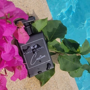 Puede incluir: Una botella cuadrada negra con una etiqueta blanca que dice "Angelic Auras Custom". La botella est&aacute; sobre una superficie marr&oacute;n claro con flores rosas y hojas verdes.